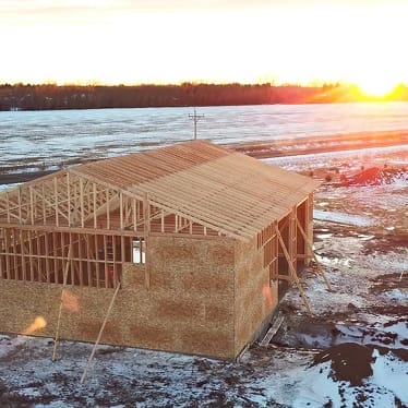 A wooden building under construction stands on snowy ground at sunset, with an open roof frame and surrounding fields in the background.