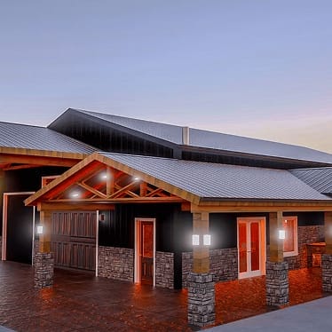 A modern building with a metal roof, stone and wood accents, illuminated exterior lights, and wide entrances at dusk.